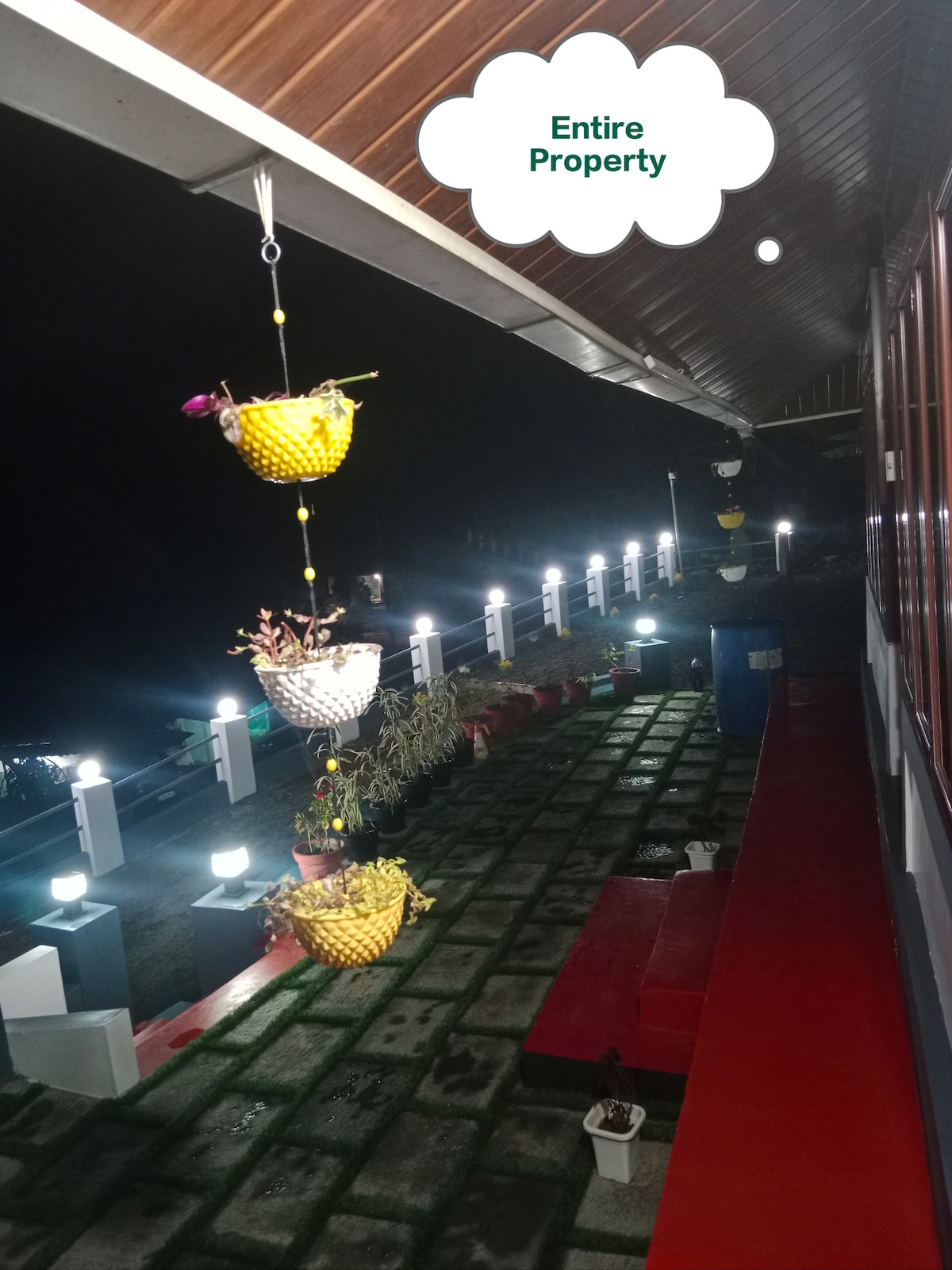 The exterior area is illuminated at night, showcasing a vibrant pathway adorned with potted plants hanging from a roof overhang. A red seating area is visible alongside the pathway, complemented by outdoor lighting that enhances the serene ambiance.