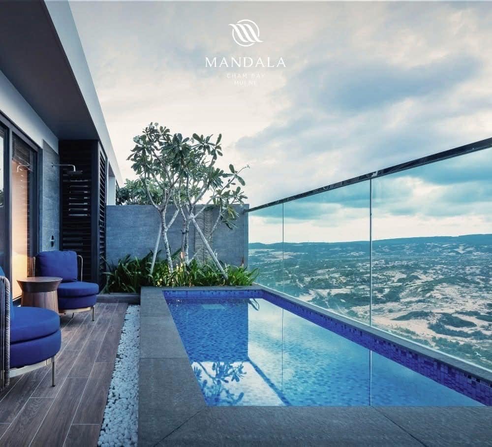An outdoor terrace features a private pool with a panoramic view. Two round chairs and a small table are positioned beside the pool, surrounded by lush greenery and smooth pebbles. The distant landscape is visible against a partially cloudy sky.