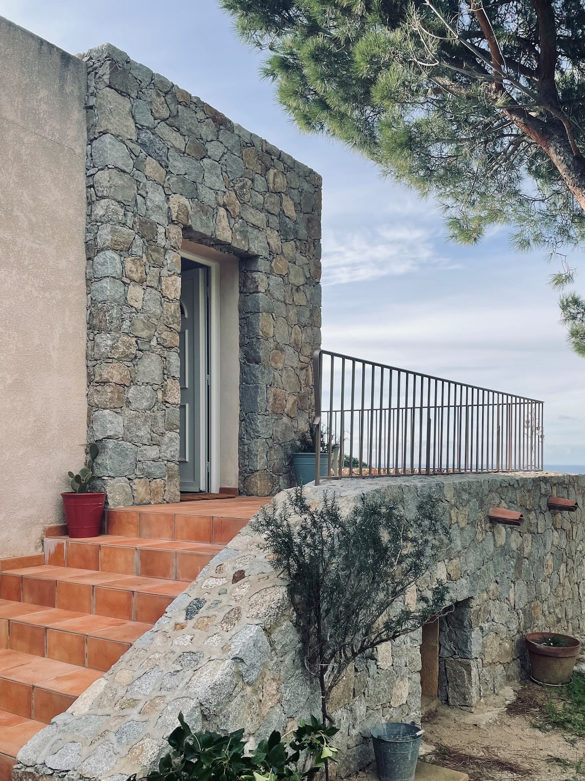 A stone-clad exterior is showcased, featuring a set of terracotta steps leading up to a doorway. A railing provides safety along a small landing, with potted plants positioned on the side. A tree stands nearby, enhancing the natural ambiance.