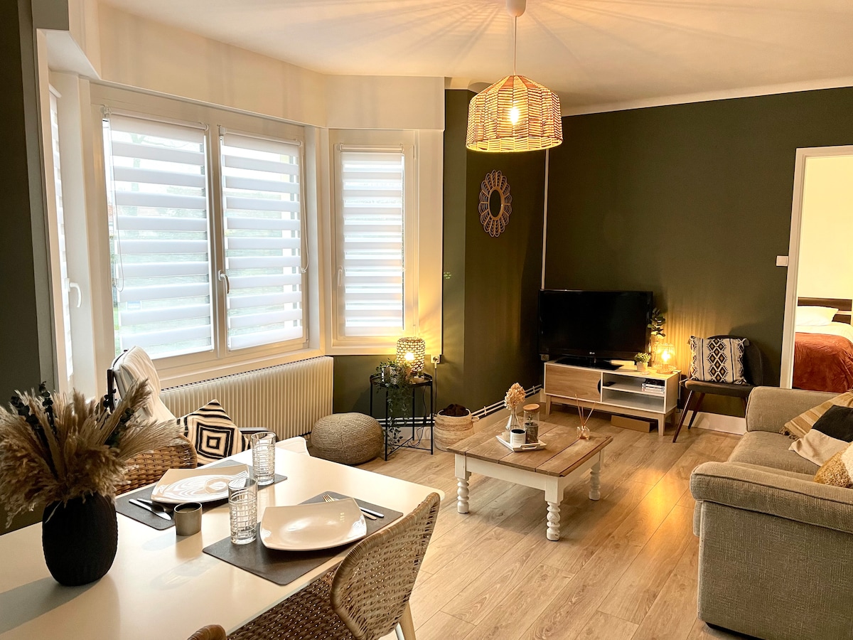 The living area showcases a blend of comfort and style with a spacious sofa and a wooden coffee table. Light filters through the slatted windows, illuminating a neutral color palette. Decorative elements include a woven pendant light and plants, creating a cozy atmosphere.