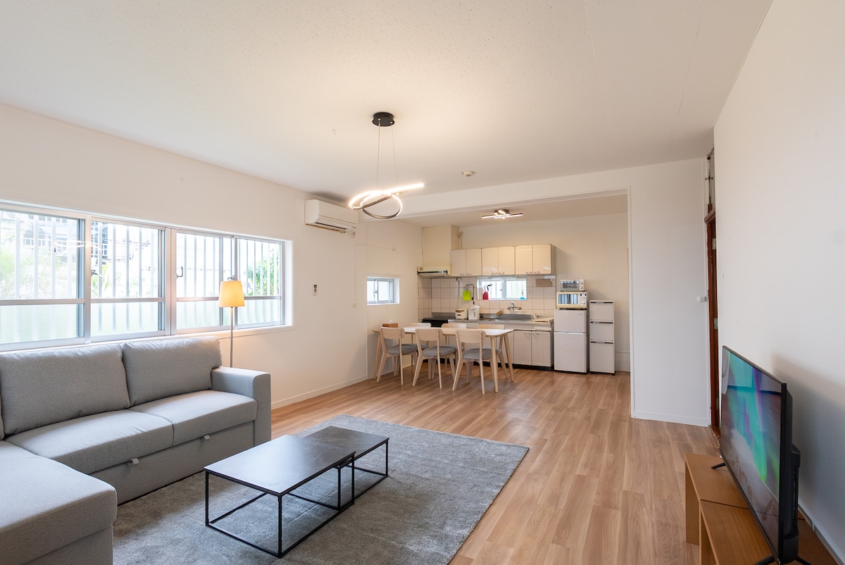 A bright living area features a light gray sofa facing a low coffee table and a flat-screen TV on a stand. A dining area, equipped with a wooden table and chairs, is visible in the background next to a modern kitchen with white cabinets.