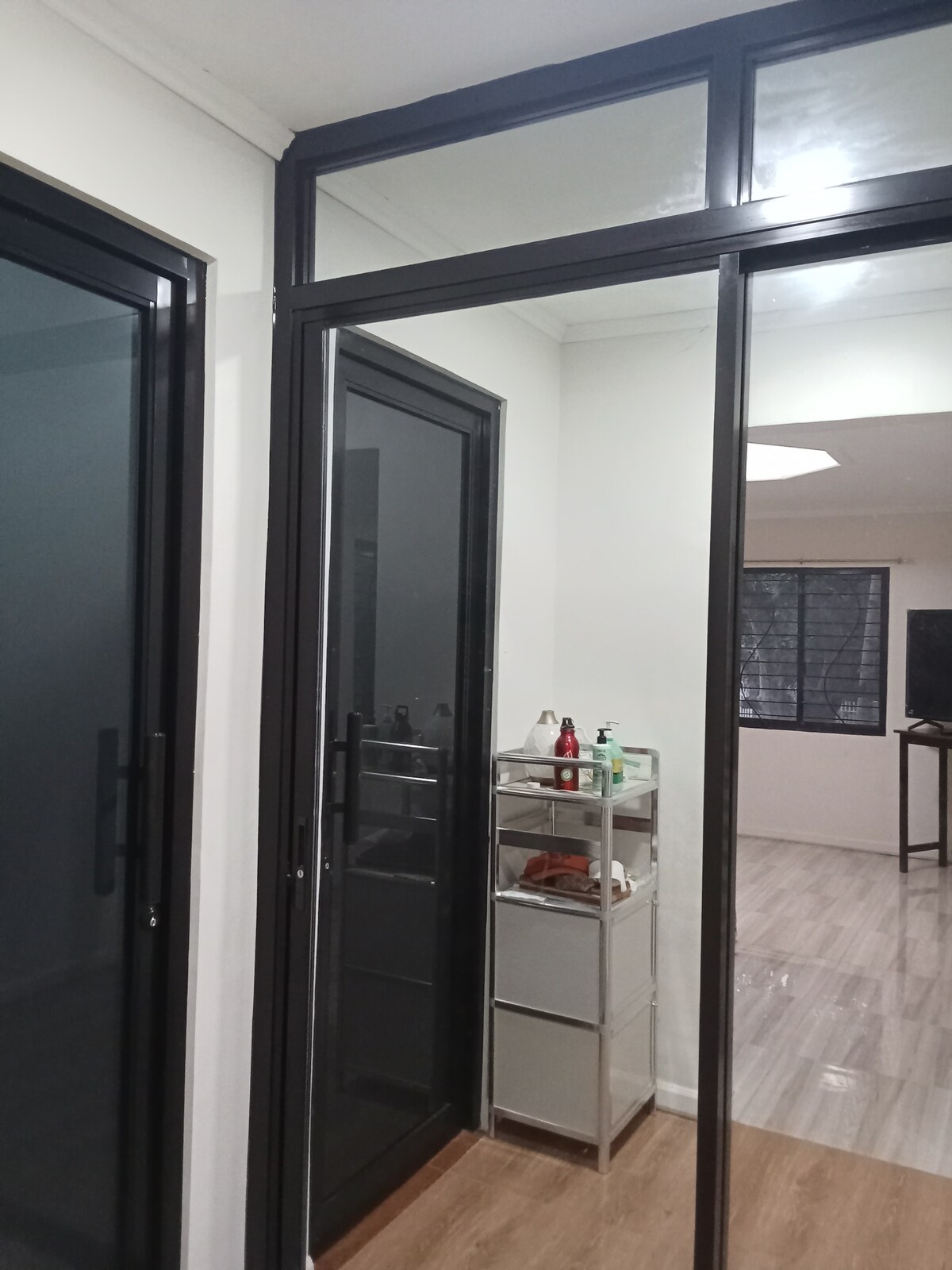 A corridor is shown, featuring sleek black framed doors leading into adjacent rooms. A small shelving unit is positioned against the wall, displaying various personal items. Natural light filters in through a nearby window, illuminating the modern design of the space.