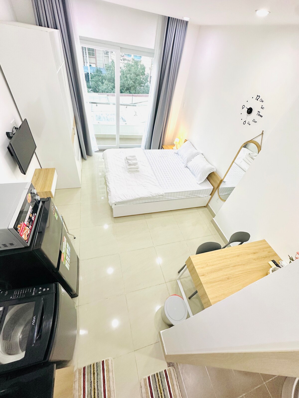 A bright and airy studio is showcased, featuring a spacious bed with white linens. A small dining area with two chairs sits adjacent to a well-equipped kitchenette. Natural light streams through large windows, and soft, neutral tones create a calm environment.