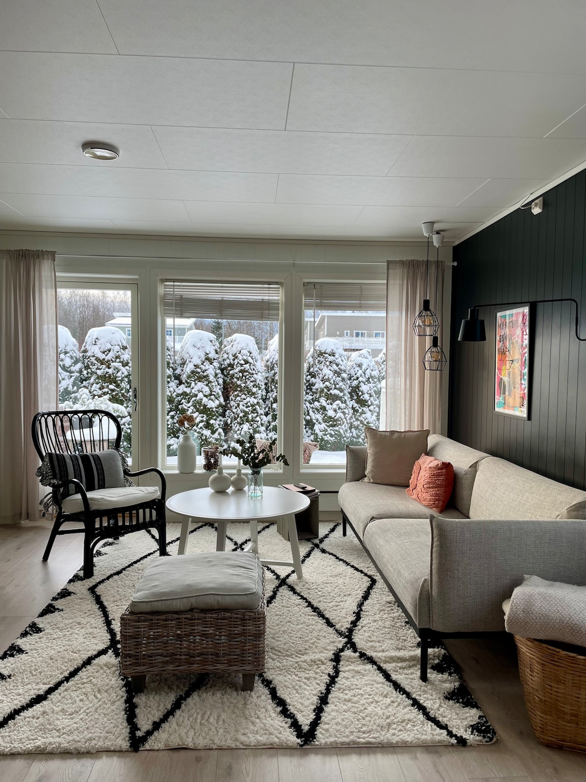 A comfortable living area is highlighted by a light-colored sofa complemented by various decorative cushions. A small round table is positioned on a patterned rug, alongside a stylish armchair. Snowy views are visible through large windows, framed by soft curtains.