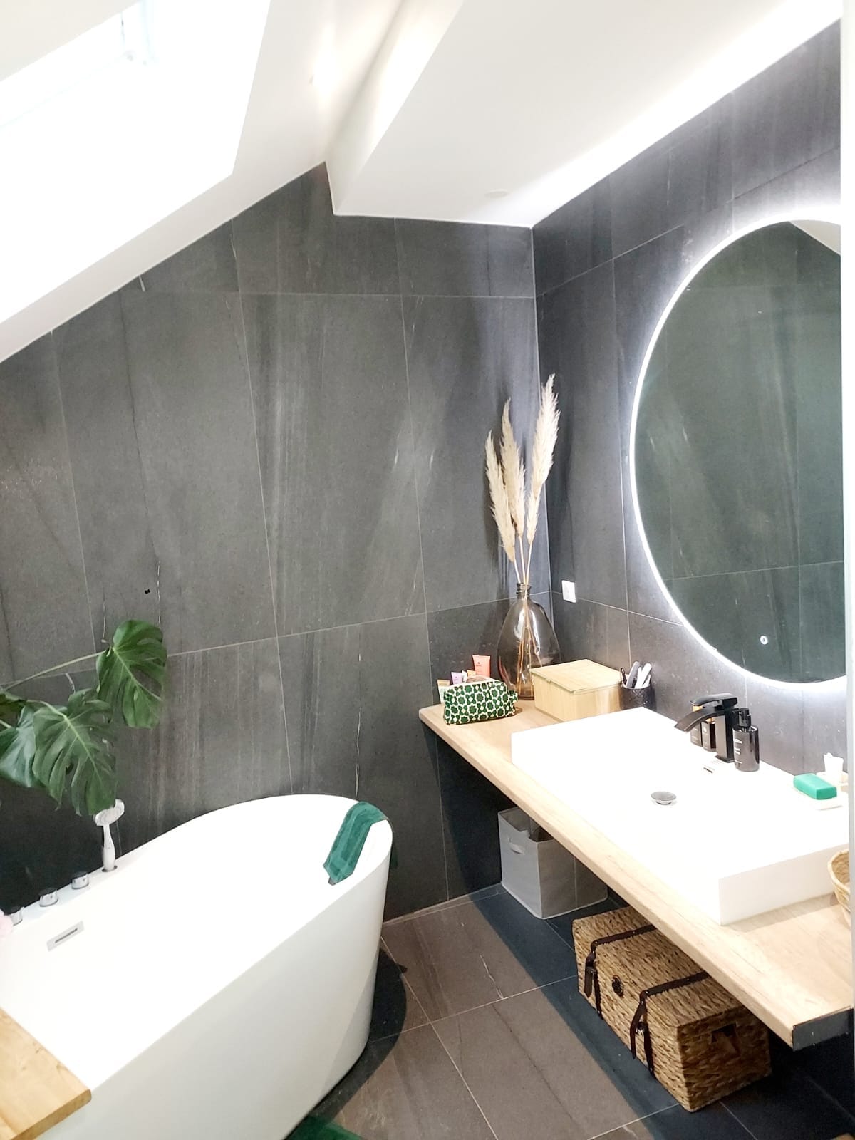 A modern bathroom features a freestanding tub alongside a wooden vanity, illuminated by soft lighting. Natural elements are included with a potted plant and dried grasses in a vase. A large round mirror complements the neutral tones of the sleek tiled walls.