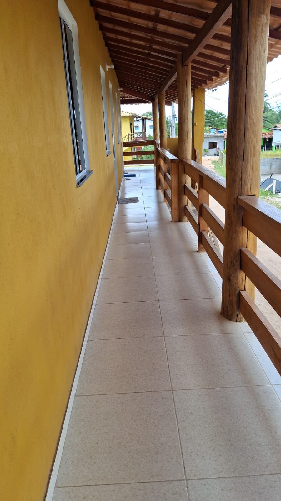 A long corridor featuring a smooth tiled floor is bordered by a vibrant yellow wall. Wooden railings frame the walkway, which is partially shaded by a rustic overhead structure. Natural light is welcomed through windows along the side, providing a bright and airy feel.