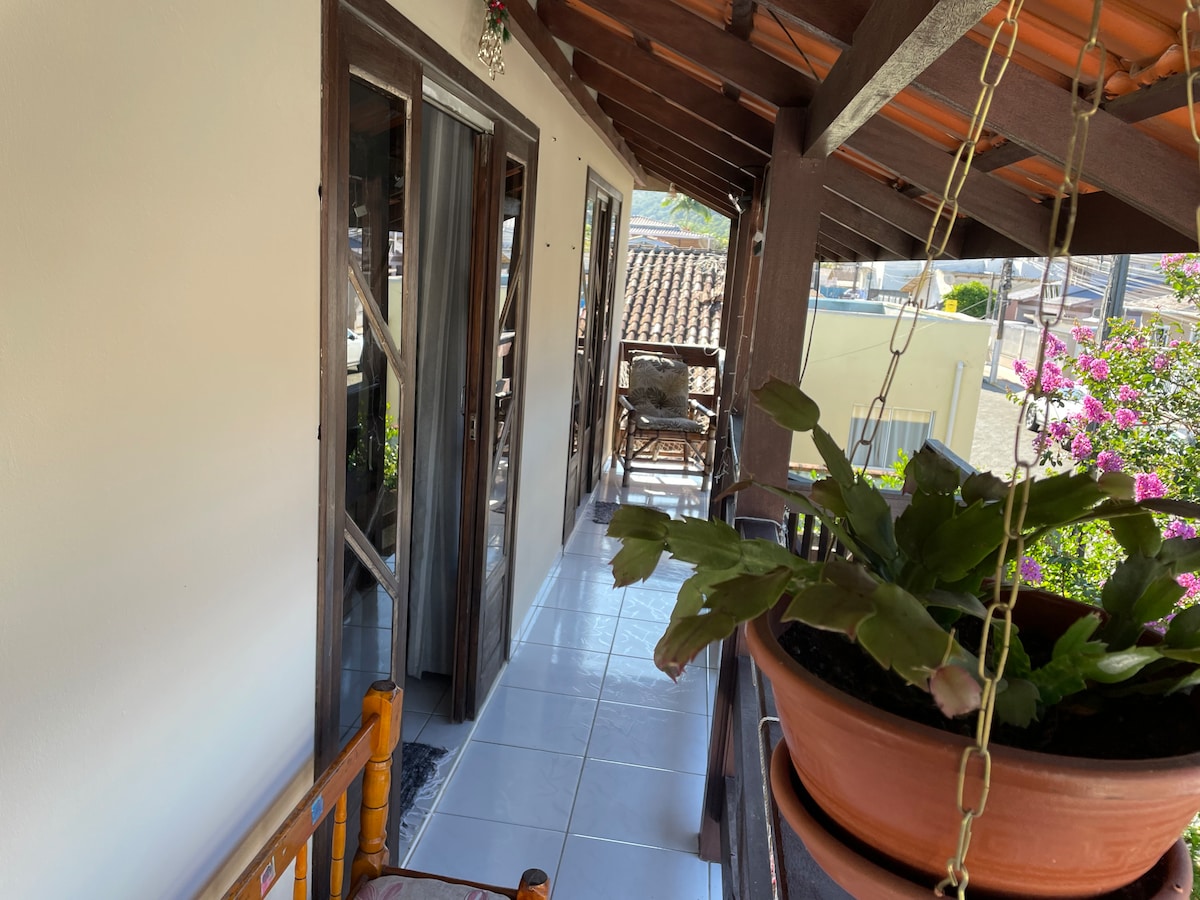 A covered balcony is visible, featuring a tiled floor and a slight glimpse of the surrounding landscape. Potted plants hang from the ceiling, while wooden furniture is placed along the porch. Open doorways lead to two rooms, adding to the open and airy feel of the space.