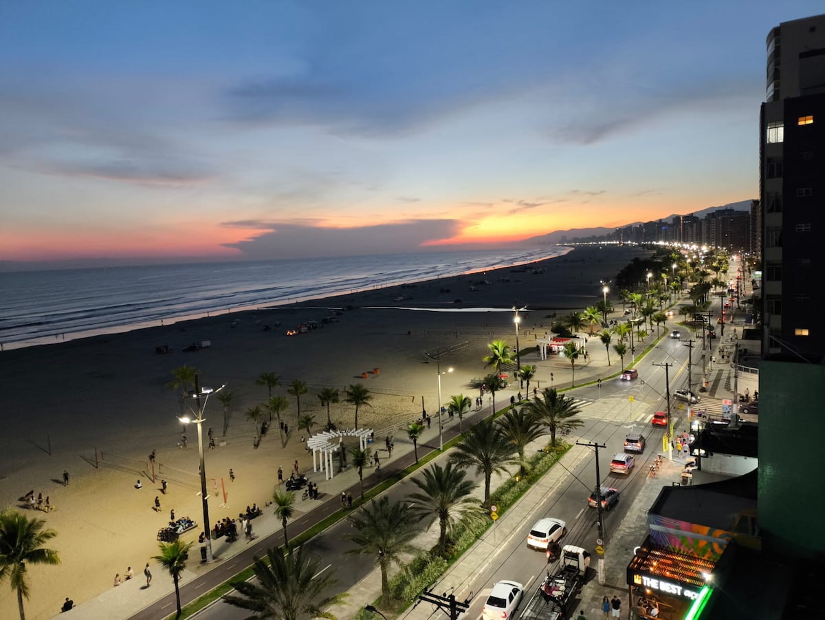 A sunset view illustrates a beachside promenade lined with palm trees. The ocean's gentle waves reflect hues of orange and pink, while people can be seen enjoying the sandy beach. Lights illuminate the pathway, creating a relaxed atmosphere along the waterfront.