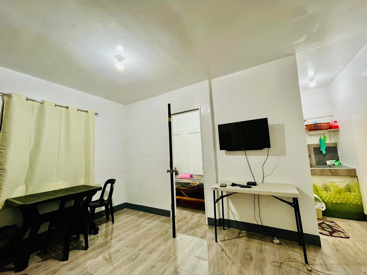 An open living area is presented with light-colored walls and sleek tiled flooring. A dark wooden table with chairs occupies one side, while a wall-mounted TV is fixed opposite. A doorway leads to a bedroom visible in the background, with soft lighting illuminating the space.