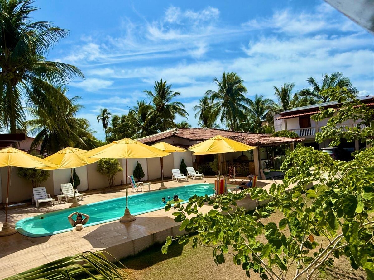 A refreshing swimming pool is surrounded by vibrant yellow umbrellas providing shade. Lounge chairs are positioned along the poolside, set against a backdrop of lush palm trees and a well-maintained garden. The structure in the background houses casual seating areas for relaxation.