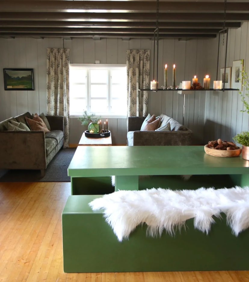 A comfortable living area features two sofas placed opposite each other, with soft cushions for added comfort. A green table with a furry throw rests in the center, surrounded by candles and decorative items. Natural light enters through a window adorned with simple curtains.