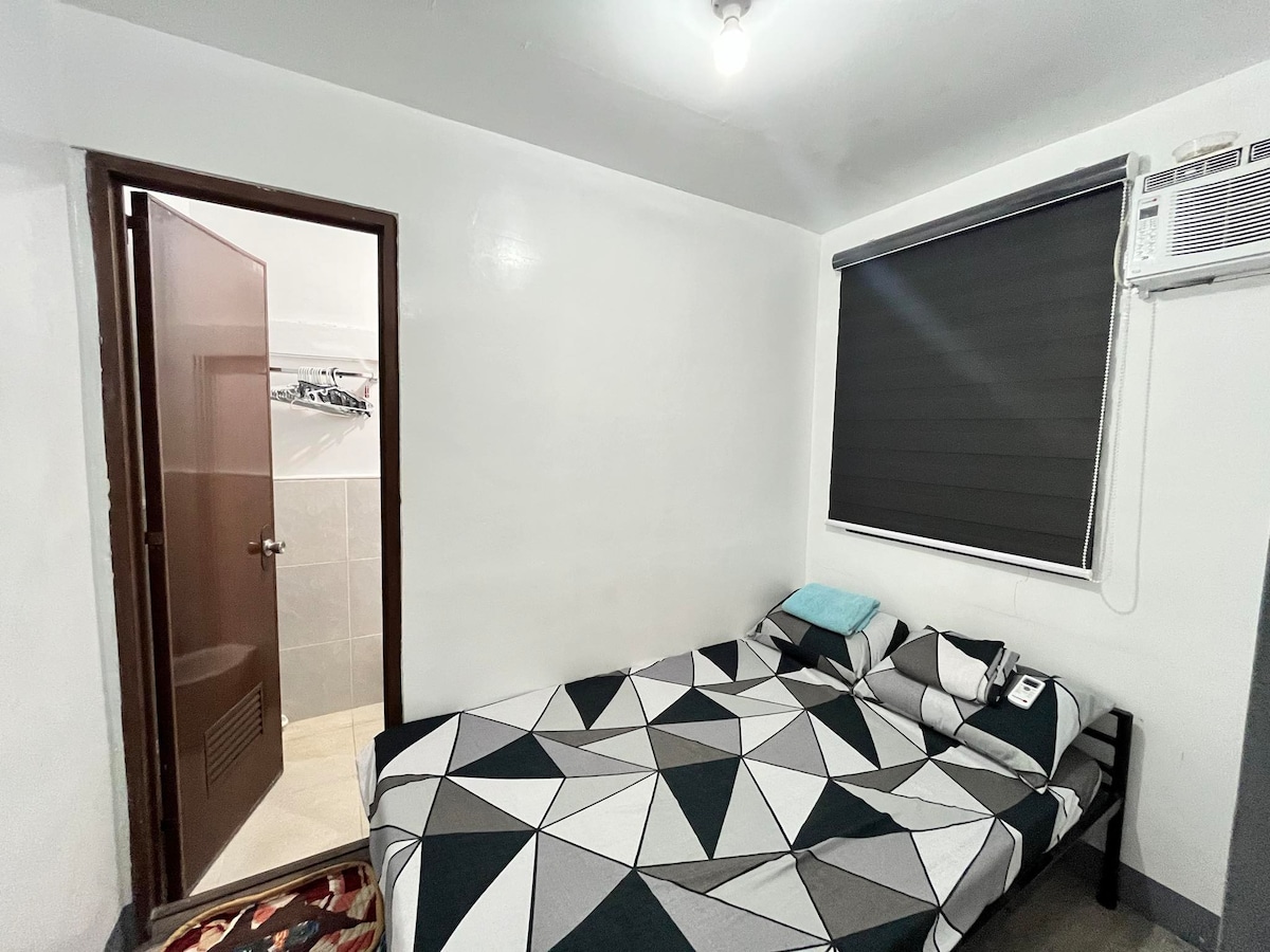 A tidy bedroom features a spacious bed with a geometric-patterned coverlet. A window with dark blinds allows for light control. An air conditioning unit is mounted on the wall, while a door leads to the adjacent comfort room.