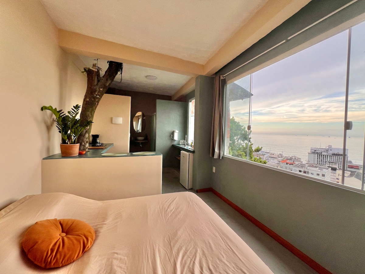 A cozy studio features a spacious bed with a round orange cushion. A small kitchenette is visible along with a mirror and sink. Large windows provide a panoramic view of the ocean, allowing natural light to fill the room, while a tree is integrated into the design.