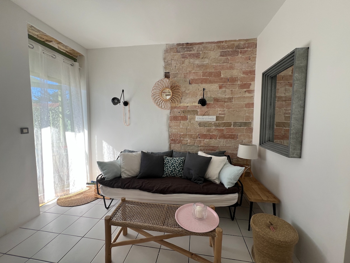 The living area features a comfortable daybed adorned with a mix of cushions in various shades. A textured stone wall serves as a backdrop, and natural light enters through sheer curtains. A wooden table and decorative items enhance the inviting space.