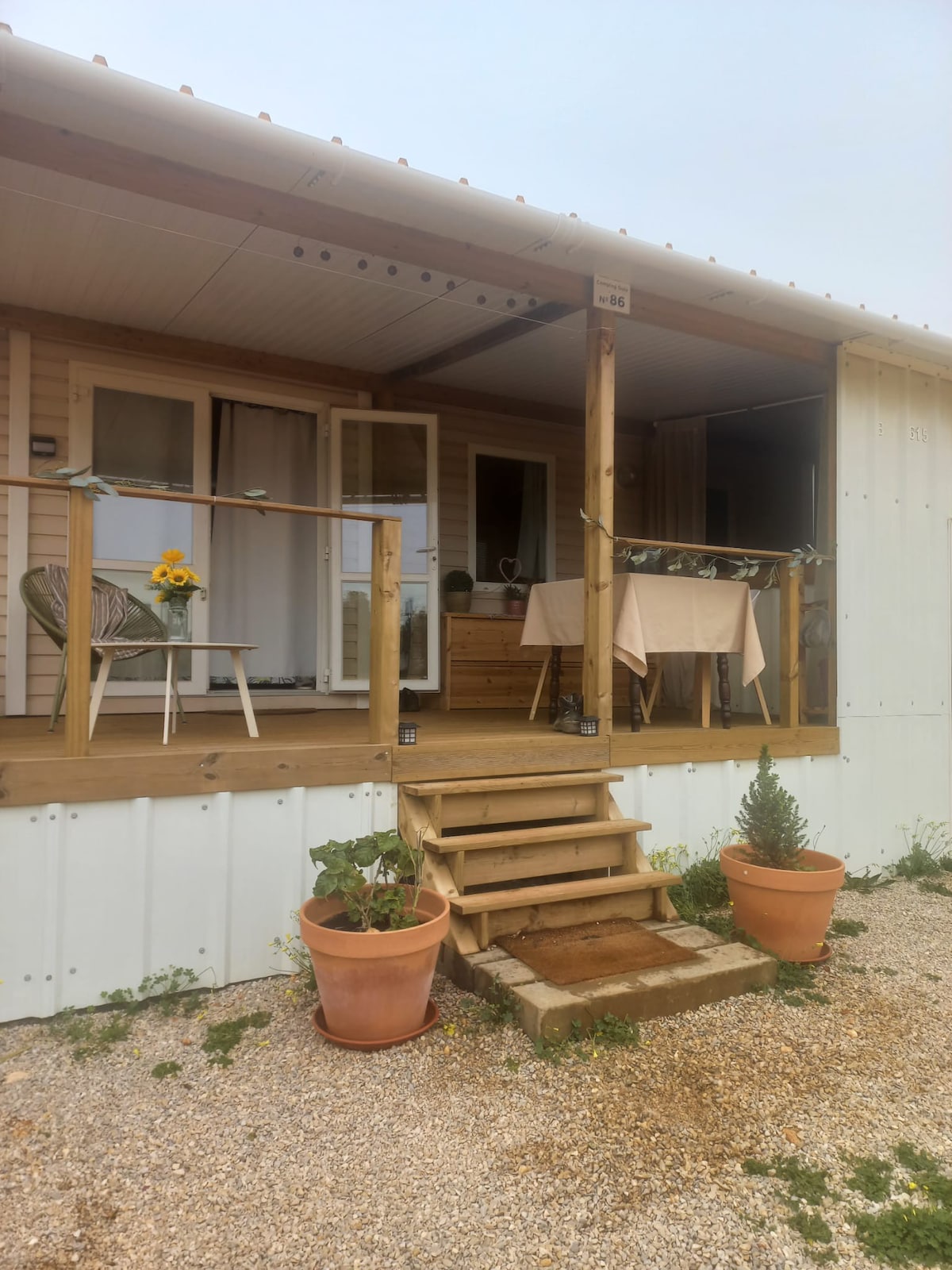 A welcoming porch features wooden steps leading to a cozy seating area. Potted plants flank the entrance, and a table is set for dining, complemented by decorative tableware. The pleasant outdoor space invites relaxation and enjoyment of the surrounding natural environment.