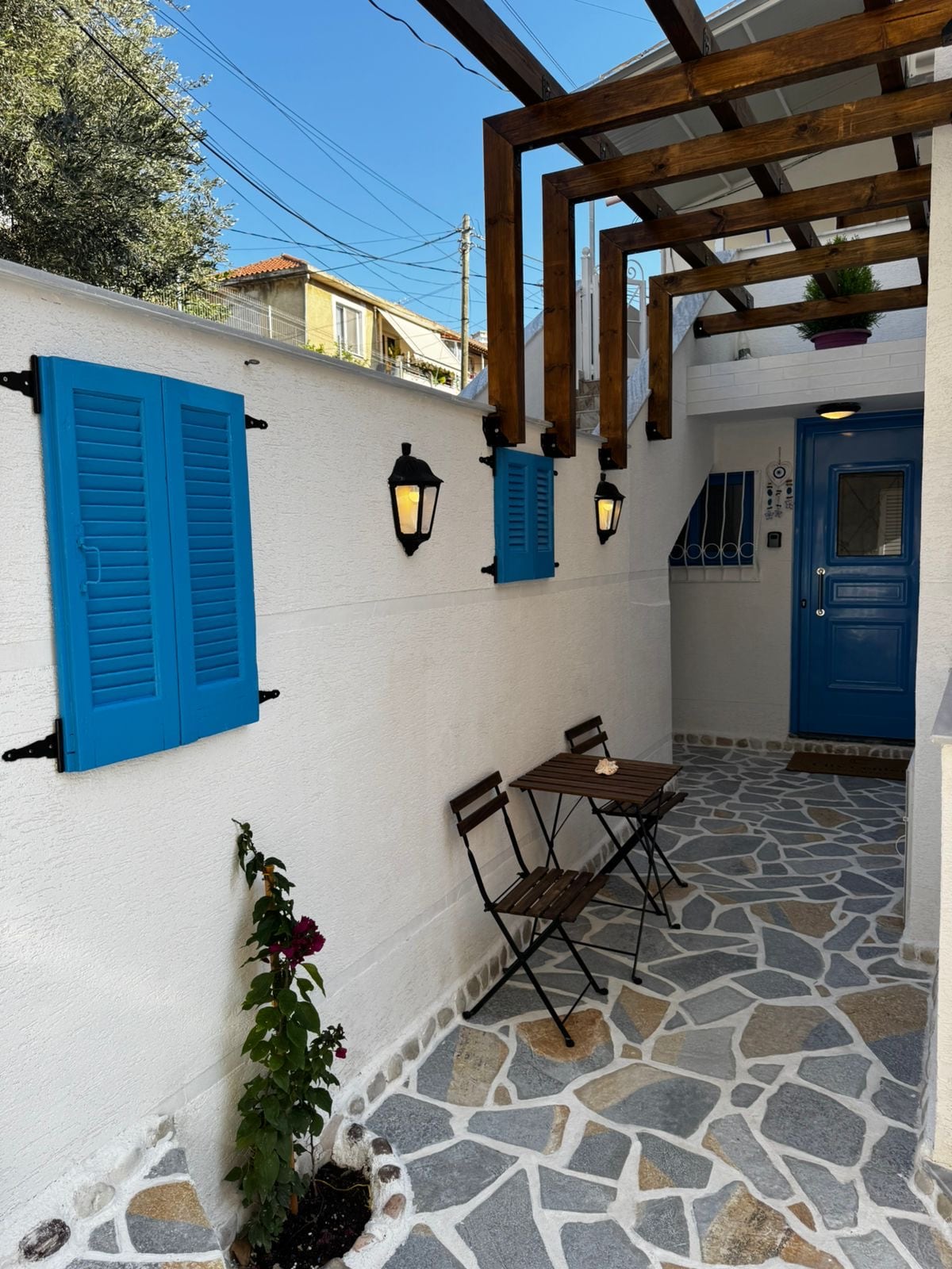 A charming outdoor area is highlighted by blue shutters and lanterns against a white wall. A small wooden table and two folding chairs are set on a patterned stone pathway, complemented by a flowering plant in a circular planter.