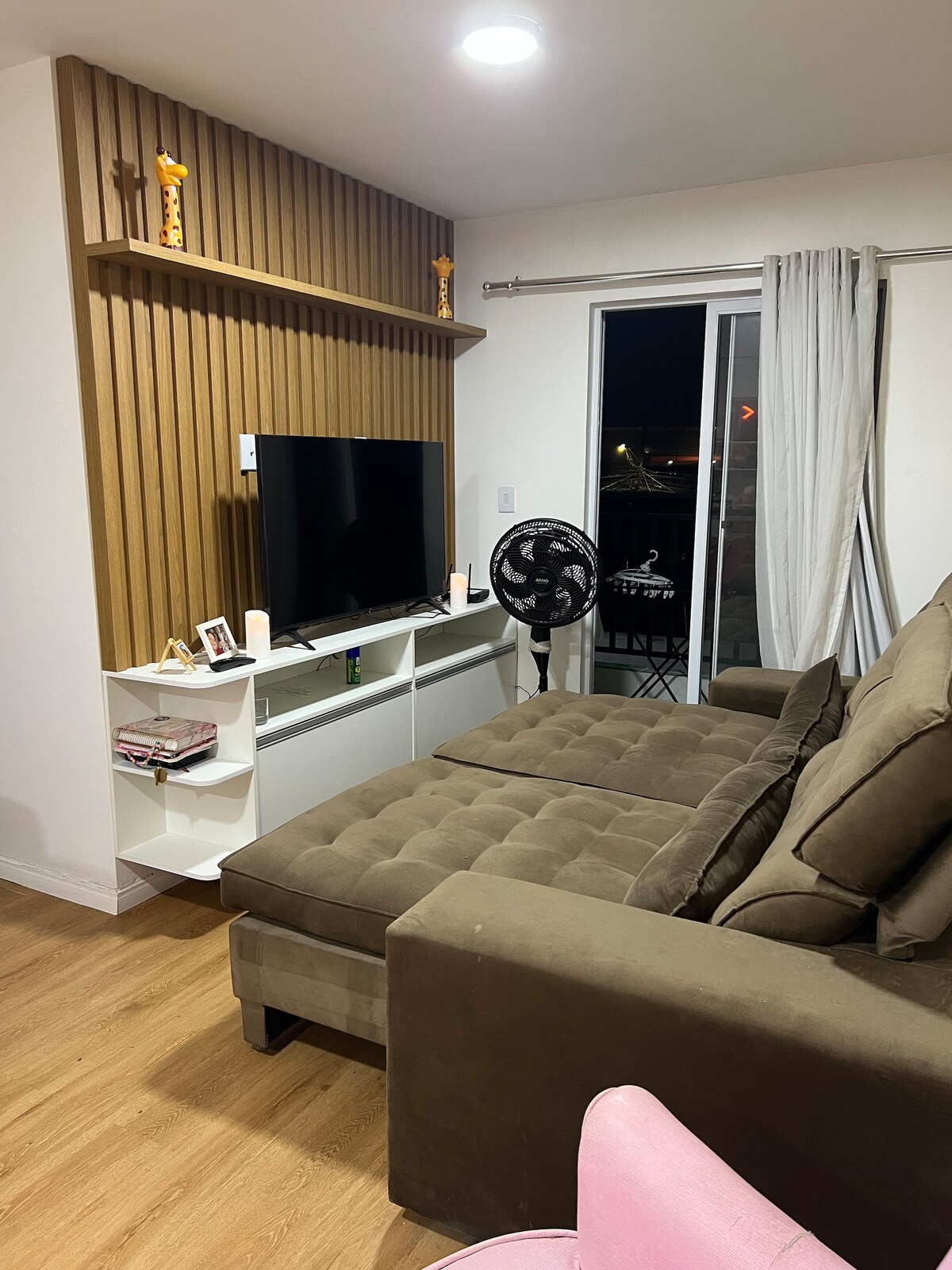 A modern living space features a large, plush sectional sofa and a sleek television mounted on a wooden accent wall. A small shelf holds decorative items, and natural light enters through a balcony door, complemented by flowing white curtains.