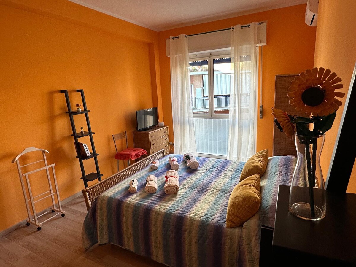 A bright room features an orange accent wall, enhancing the sense of space. A spacious bed is adorned with a multicolored blanket. A small TV, a shelf, and a dresser are visible. Light enters through a window covered by sheer curtains, providing a warm ambiance.