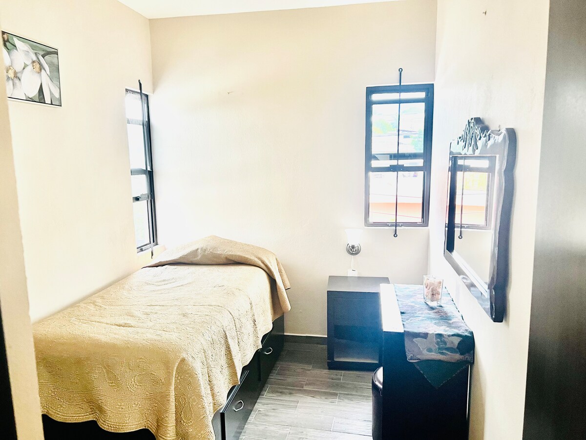 A cozy sleeping area is depicted, featuring a single bed covered with a light-colored blanket. Two windows allow natural light to fill the room, and a dark wooden bedside table complements the space. A mirror and small vanity are positioned across from the bed.