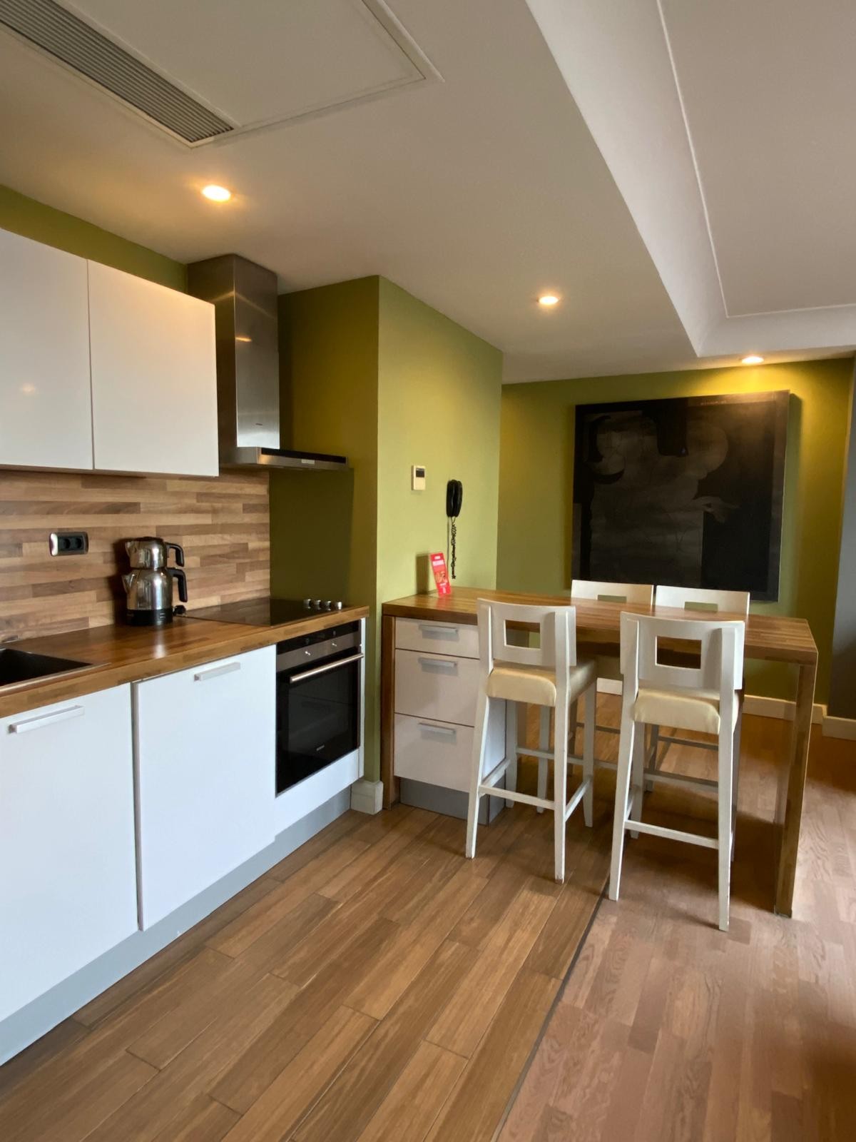 A modern kitchen space features sleek white cabinetry and a wood-paneled accent wall. Stainless steel appliances, including an oven and coffee maker, are present. Adjacent, a dining area is equipped with a wooden table and four high-backed chairs, set against a dark artwork backdrop.