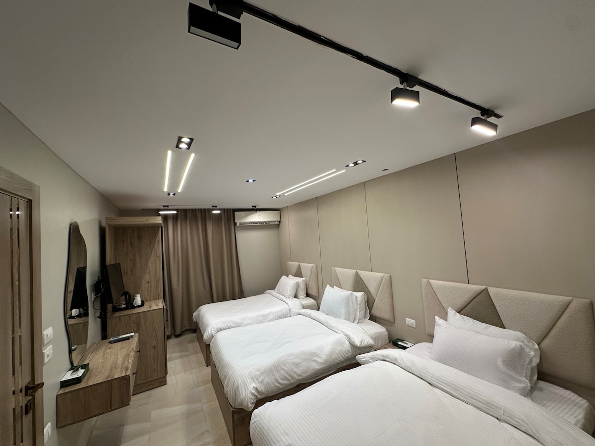 A spacious room is furnished with three single beds, featuring light bedding and soft headboards. A wooden desk is positioned against the wall, accompanied by a wall-mounted television. Warm lighting is provided by recessed fixtures, enhancing the modern design.