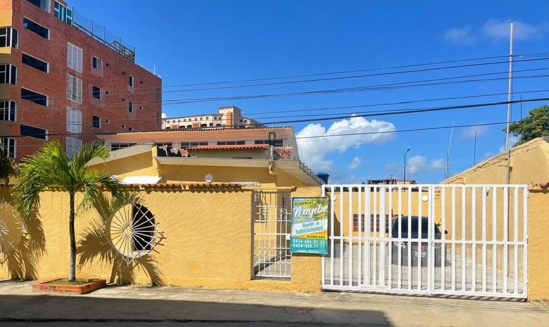 A secure entrance featuring a white gated fence with circular designs stands in front of a yellow building. The bright blue sky serves as a backdrop, complementing the vibrant colors of the property. A palm tree adds a touch of greenery to the entrance area.