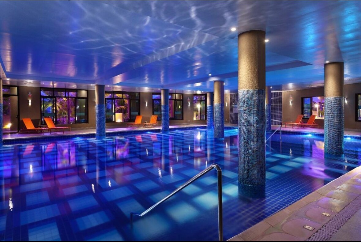 An indoor swimming pool is illuminated by colorful lights, creating a calming atmosphere. A set of orange lounge chairs is positioned around the water's edge, while pillars lined with blue tiles support the ceiling.