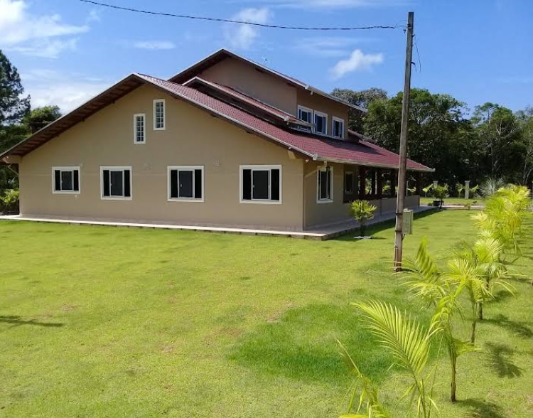 The spacious house is set against a backdrop of lush greenery, featuring a large lawn and multiple windows that allow for natural light. A red-tiled roof enhances the architectural appeal, while the surrounding landscape is well-maintained and offers a serene outdoor setting.