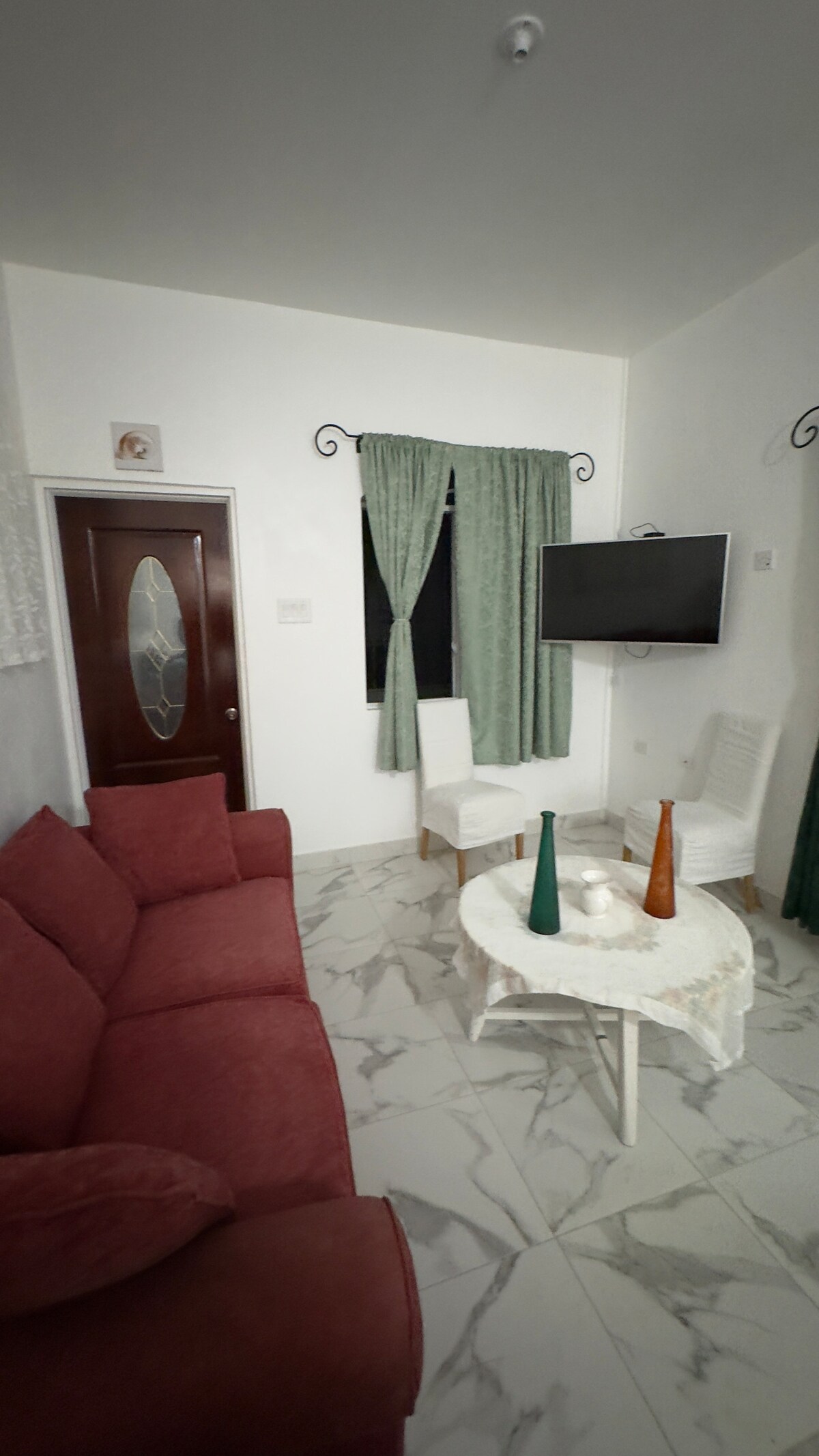 An open living area features a marbled floor with a plush red sofa and a circular coffee table at its center. Two white chairs are positioned beside the table, with a wall-mounted flat-screen TV providing entertainment. Long curtains frame the window, allowing natural light to filter in.