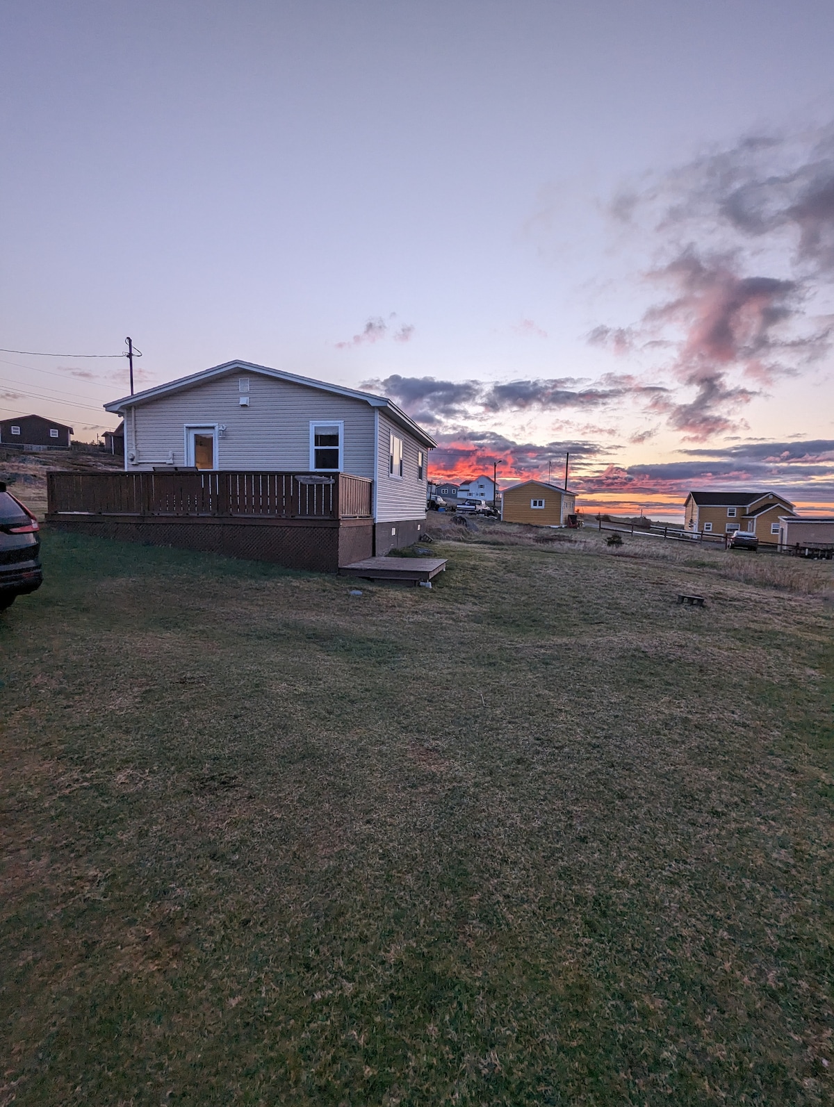 The exterior of the cozy cottage is shown against a colorful sunset sky, with clouds reflecting hues of orange and pink. A wooden deck is visible at the front, alongside a grassy area leading to the rocky shoreline. Nearby, additional cottages are outlined at dusk.