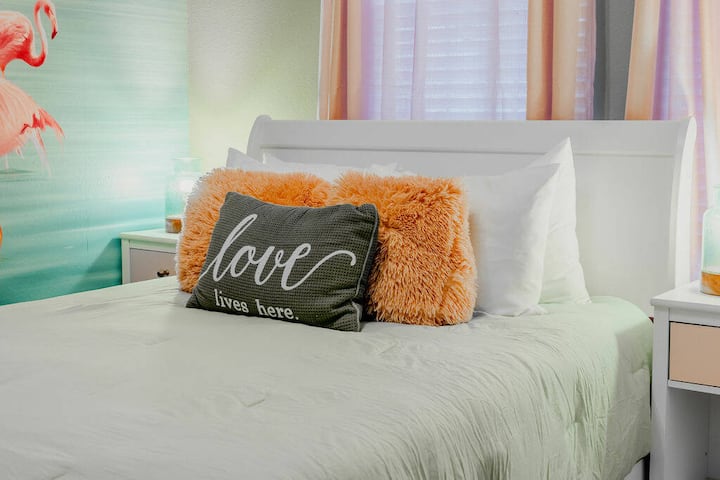 This cozy bedroom features a soft, light green color palette complemented by playful orange pillows that add a cheerful touch. The welcoming bed is flanked by sleek nightstands, making it a delightful spot to unwind and recharge.