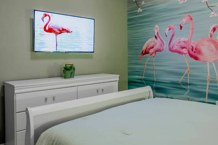 Welcome to this vibrant bedroom, where a stunning flamingo mural adds a playful touch to the serene colors. The sleek white furniture enhances the space, and the wall-mounted TV is perfect for cozy movie nights.