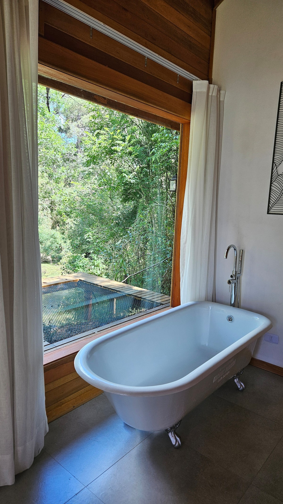 A white, freestanding bathtub is positioned near a large window, offering views of lush greenery outside. Sheer white curtains frame the window, allowing natural light to brighten the space, while a sleek faucet is mounted beside the tub.