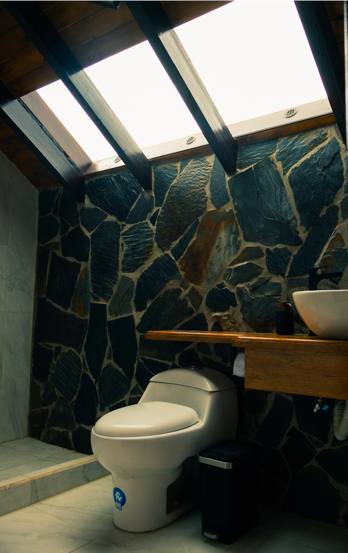 A modern bathroom features a stone wall, complemented by a wooden countertop extending over the toilet. Natural light enters through a skylight, illuminating the space. A small trash bin and a stylish basin are positioned on the countertop.