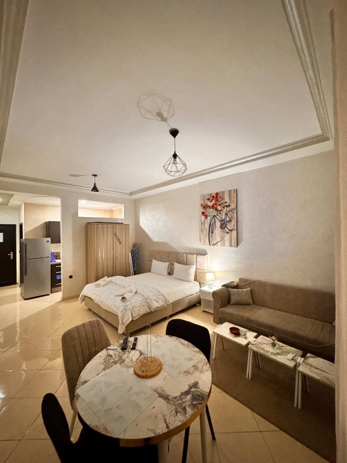 This inviting studio features a spacious bed adorned with light linen, complemented by a decorative wall piece. A comfortable sofa and a dining table are arranged, creating a cozy living area. Soft lighting and neutral tones enhance the warmth of the space.