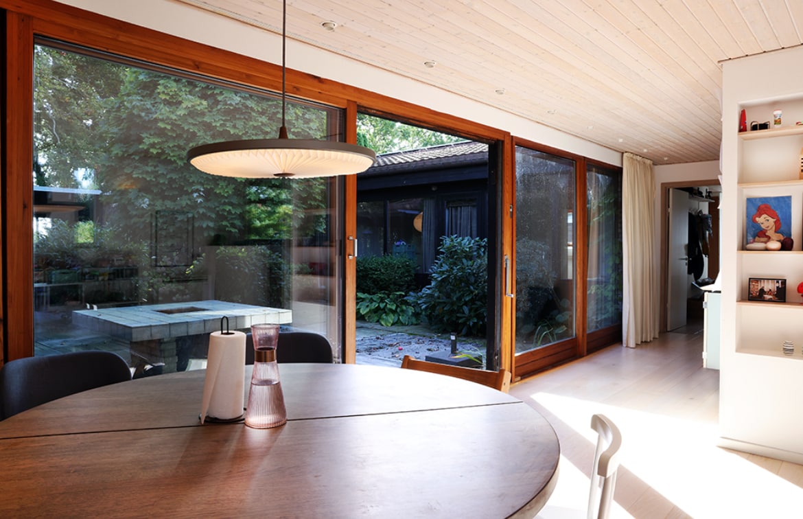 An open dining area is illuminated by natural light, featuring a round wooden table and modern chairs. Large glass doors provide a view of the private garden, where greenery complements the serene environment. A bright, airy ambiance is created by the wooden ceiling and light-colored walls.