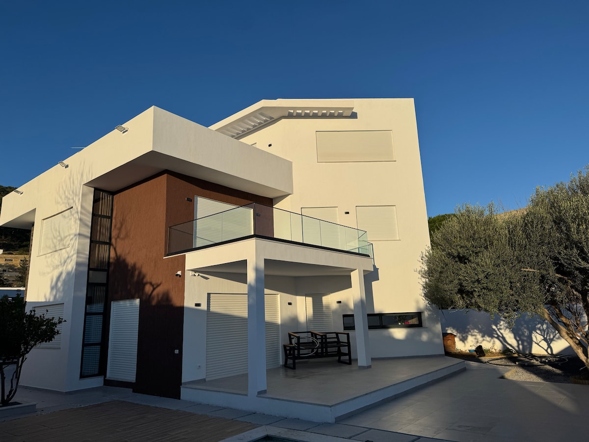 A modern, multi-story house is depicted with a sleek white façade accented by brown panels. A spacious covered terrace extends from the second floor, featuring glass railings and a dining table beneath. The surrounding area is bordered by olive trees and a clear sky.