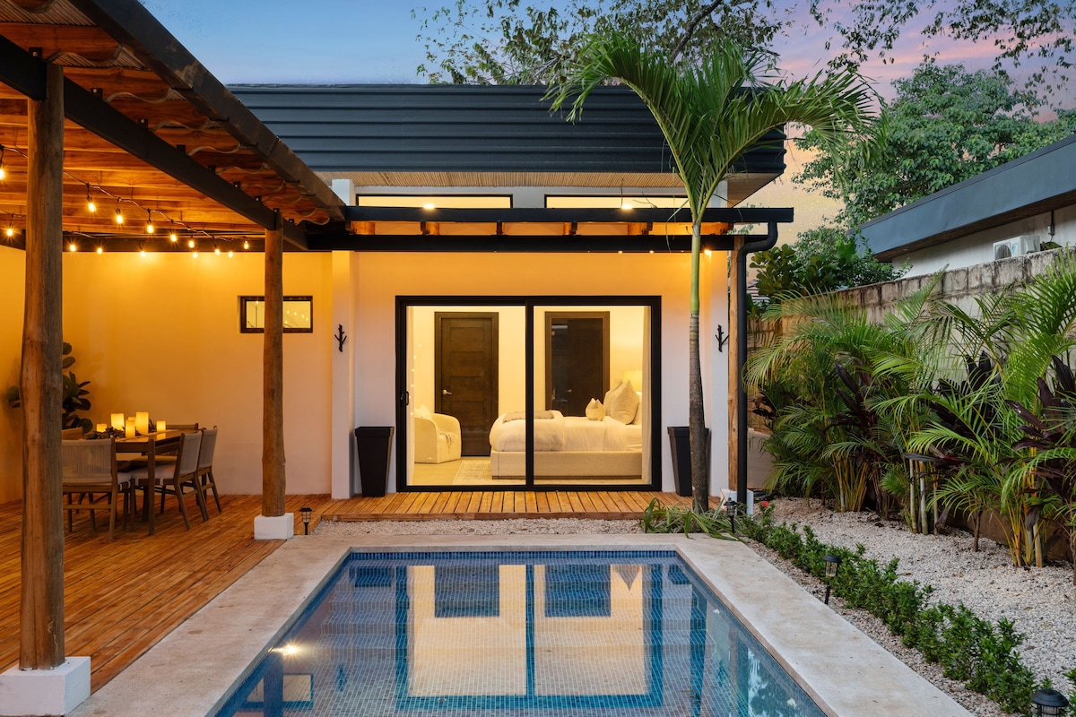 A serene outdoor area features a private pool, bordered by lush greenery, with a wooden deck extending towards sliding glass doors. Inside, a spacious bedroom is visible, allowing natural light to enhance the beautiful surroundings during sunset.