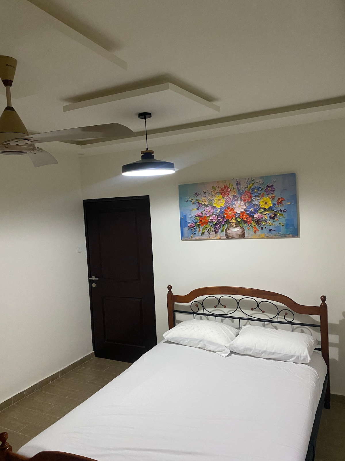 A serene bedroom is equipped with a single bed dressed in crisp white linens. A ceiling fan is mounted above, and a pendant light offers soft illumination. A colorful floral painting adorns the wall, adding a touch of vibrancy to the calm interior.