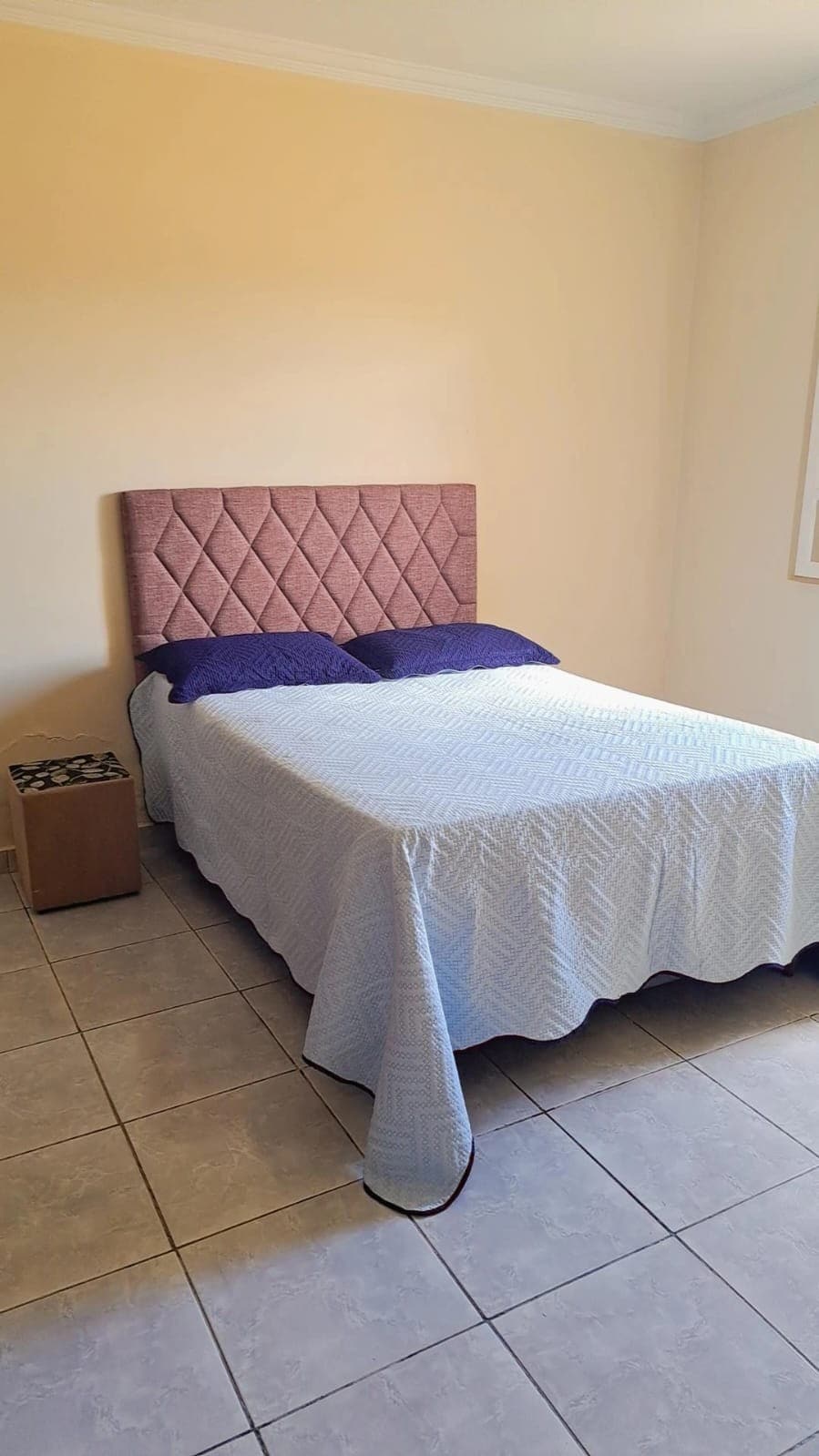 A bright bedroom features a spacious bed with a textured headboard and a light-colored quilt draped over it. A small bedside table stands beside the bed, complementing the neutral wall color and tile flooring.