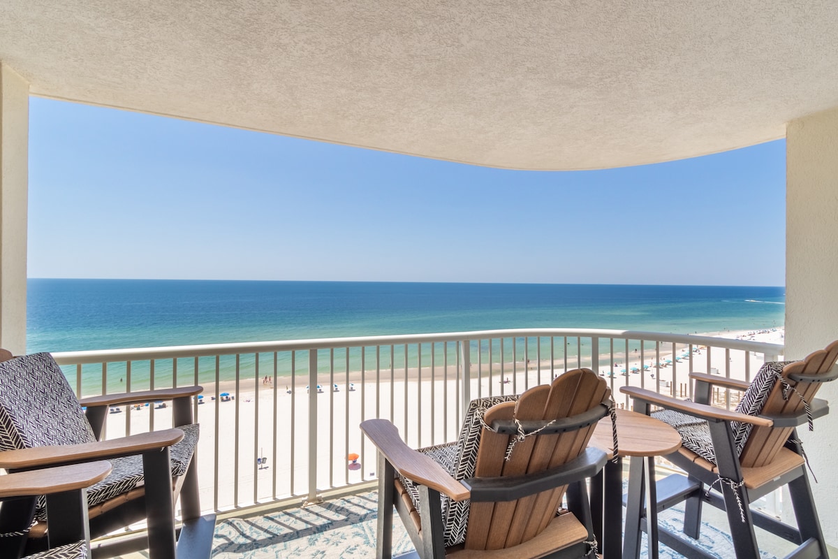 A spacious balcony overlooks a sandy beach and the ocean, featuring comfortable seating with two chairs and a table, offering a place to relax. The expansive view reveals clear blue waters meeting the horizon under a bright sky, enhancing the outdoor experience.
