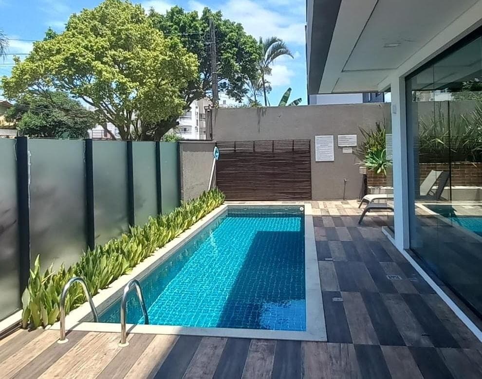A serene outdoor area features a rectangular pool framed by wooden decking and lush greenery. Clear glass panels provide privacy while allowing sunlight to enhance the tranquil environment. This space is designed for relaxation within a peaceful setting.
