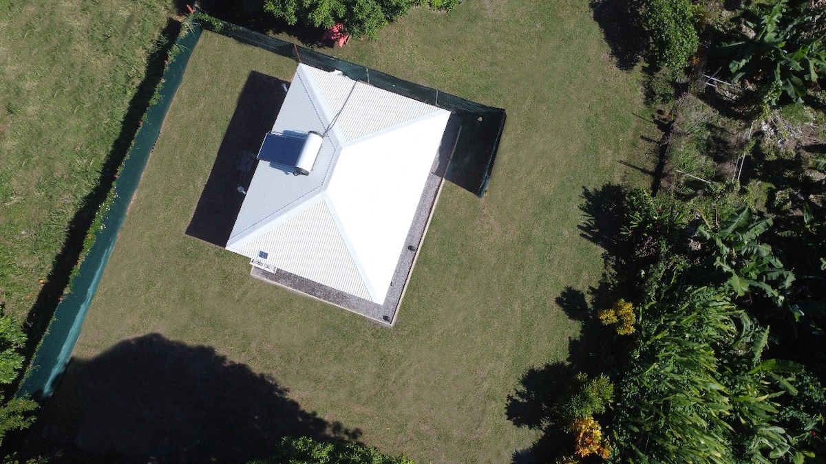 An aerial view of a bungalow surrounded by a green perimeter and a spacious lawn. The rooftop, with a distinctive shape, is visible along with elements of the surrounding garden featuring tree foliage and a fenced area.