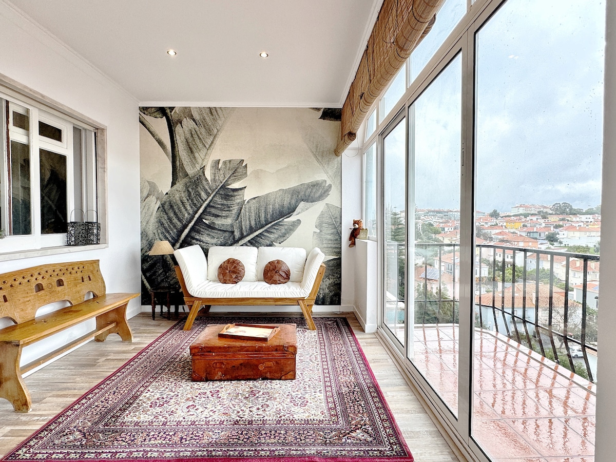 A light-filled living area features a comfortable sofa adorned with decorative pillows, complemented by a wooden bench. A rich area rug anchors the space, while large windows provide sweeping views of the cityscape, enhancing the artistic mural on the wall.