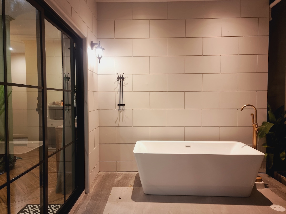 A modern bathroom features a freestanding bathtub against a textured wall. Soft light is provided by a wall-mounted fixture, and glass doors offer a view of the adjoining space. Textured tiles add warmth to the floor, complemented by greenery in the corner.