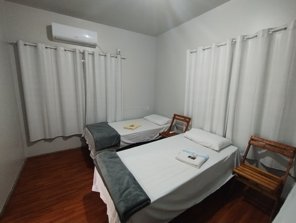 A room is furnished with two individual beds, each covered with simple white linens. A stack of towels is placed on one bed. Two wooden chairs are positioned against the wall, and light gray curtains cover the windows, allowing soft light to enter the space.