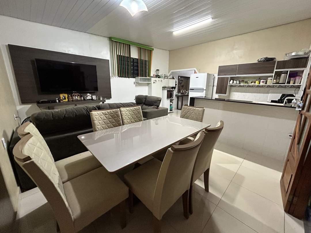 A modern dining area is showcased with a light-colored table and six upholstered chairs. A comfortable sofa is positioned nearby, and a large flat-screen television is mounted on the wall. Kitchen appliances and cabinetry are visible in the background, providing an open layout.