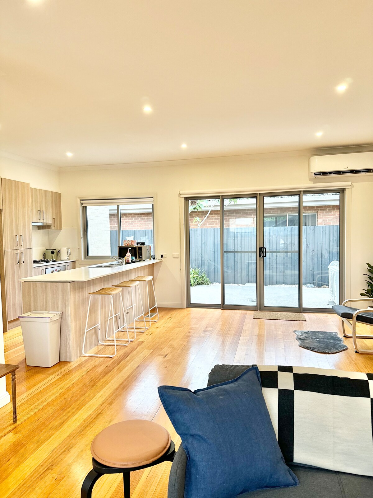An open-plan living area features wooden flooring and large windows that allow natural light to enter. A modern kitchen is visible, equipped with light wood cabinetry and bar stools. A cozy seating area is arranged with cushions and a small accent rug, creating a comfortable atmosphere.
