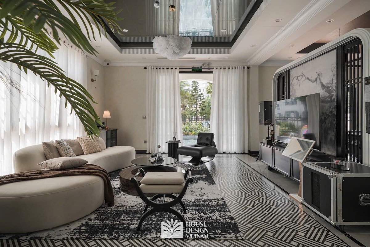A modern living area is presented with a curved sofa, a patterned rug, and a coffee table. Tall windows allow natural light to fill the space, while greenery peeks through the sheer curtains. A television is positioned on a sleek cabinet, enhancing the entertainment options.