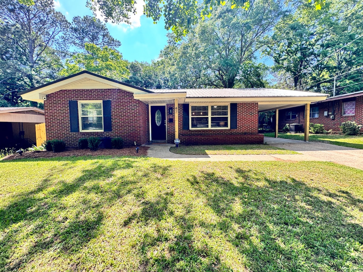 The exterior of a single-story brick home is presented, featuring a welcoming front entrance and landscaped yard. Sunlight filters through the trees, creating dappled shadows on the well-kept lawn, while a covered porch provides a shaded space for relaxation.